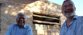 Zwei Männer sitzen lachend nebeneinander auf Holzbänken.