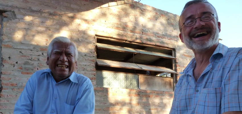 Zwei Männer sitzen lachend nebeneinander auf Holzbänken.