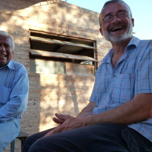 Zwei Männer sitzen lachend nebeneinander auf Holzbänken.