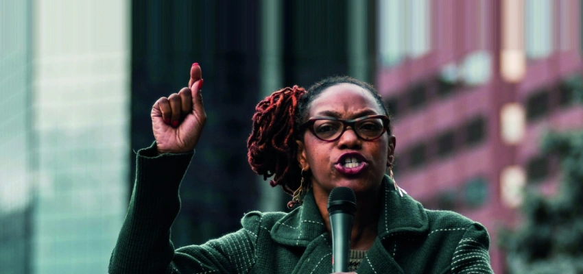 Eine Frau steht mit Mikrofon in der Hand auf einer Bühne in einer Großstadt.