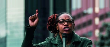 Eine Frau steht mit Mikrofon in der Hand auf einer Bühne in einer Großstadt.