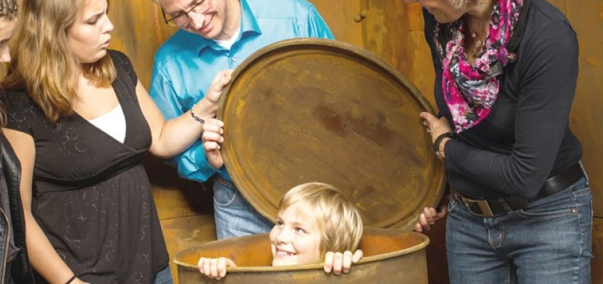 Einige Erwachsene stehen um eine Tonne, aus der, grinsend, ein Kind schaut.