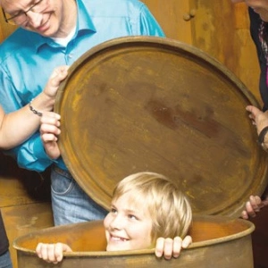 Einige Erwachsene stehen um eine Tonne, aus der, grinsend, ein Kind schaut.