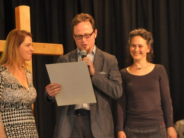 Ein Mann und zwei Frauen stehen auf einer Bühne vor einem Kreuz. Der Mann liest etwas vor.