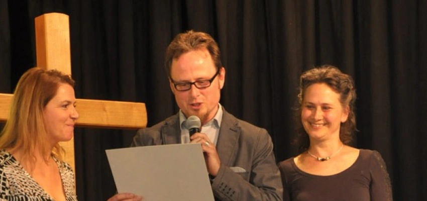 Ein Mann und zwei Frauen stehen auf einer Bühne vor einem Kreuz. Der Mann liest etwas vor.