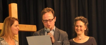 Ein Mann und zwei Frauen stehen auf einer Bühne vor einem Kreuz. Der Mann liest etwas vor.