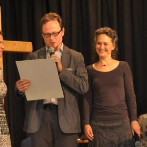 Ein Mann und zwei Frauen stehen auf einer Bühne vor einem Kreuz. Der Mann liest etwas vor.