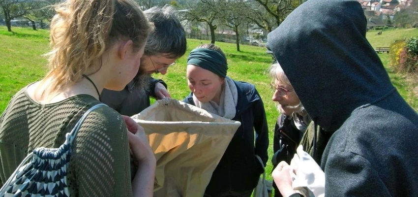 5 Personen gucken neugierig in einen dreieckigen Sack auf einer Wiese auf einem Hügel nahe eines Dorfes