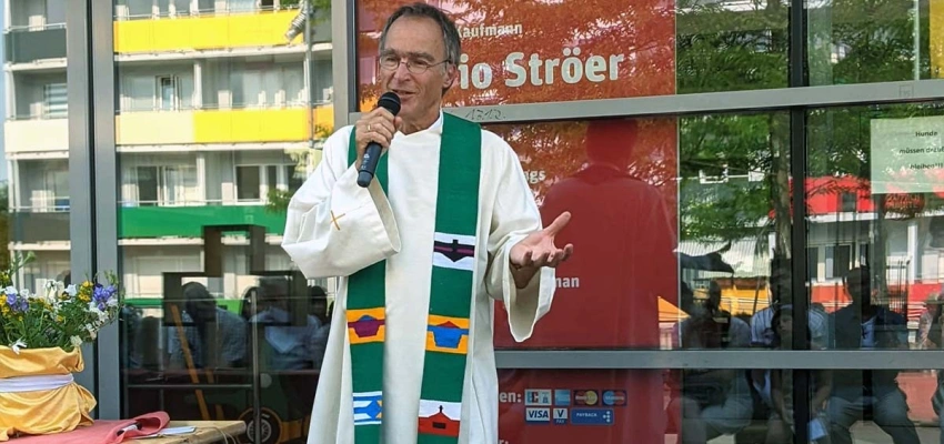 Älterer Mann mit Mkirphon und Robe spricht zu sitzenden Publikum bei Sonnenschein vor einem Kaufmann in der Stadt