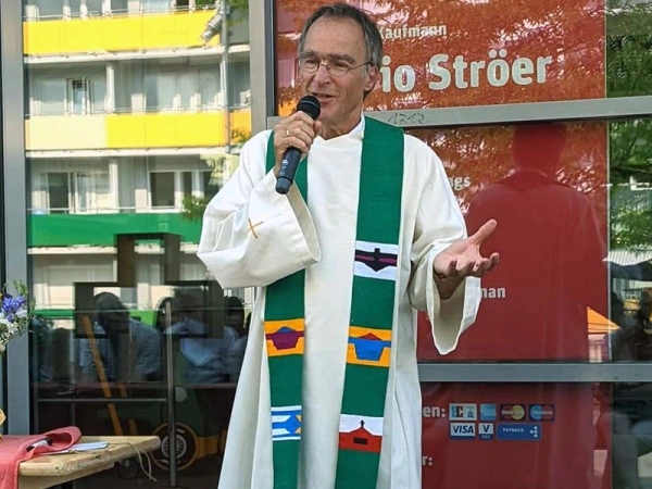 Älterer Mann mit Mkirphon und Robe spricht zu sitzenden Publikum bei Sonnenschein vor einem Kaufmann in der Stadt