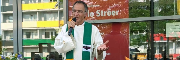 Älterer Mann mit Mkirphon und Robe spricht zu sitzenden Publikum bei Sonnenschein vor einem Kaufmann in der Stadt