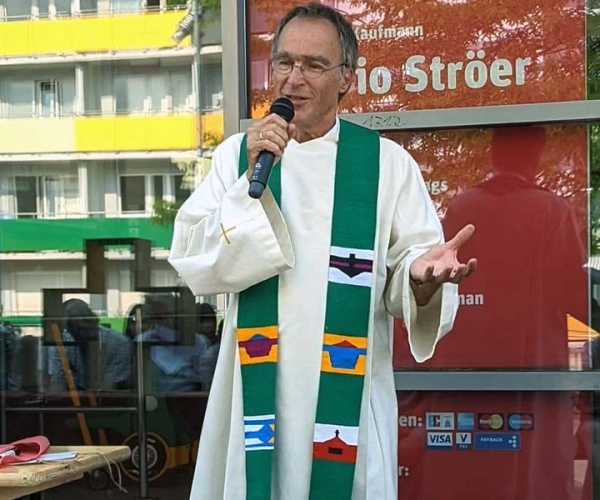 Älterer Mann mit Mkirphon und Robe spricht zu sitzenden Publikum bei Sonnenschein vor einem Kaufmann in der Stadt