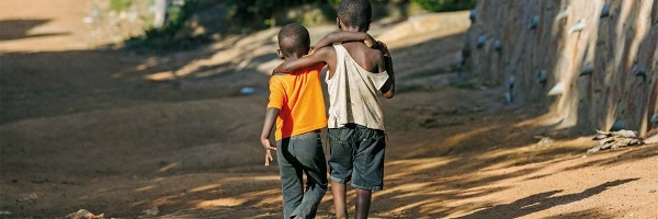 Zwei schwarze, barfüßig gehende, Kinder, Schulter an Schulter auf einem Sandweg